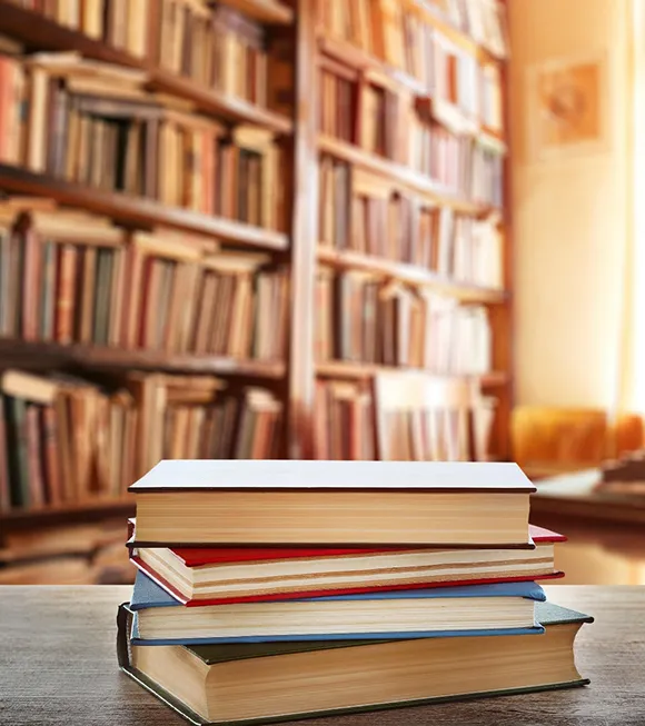Stack of books on a wooden table, with bookshelves filled with more books in the background.